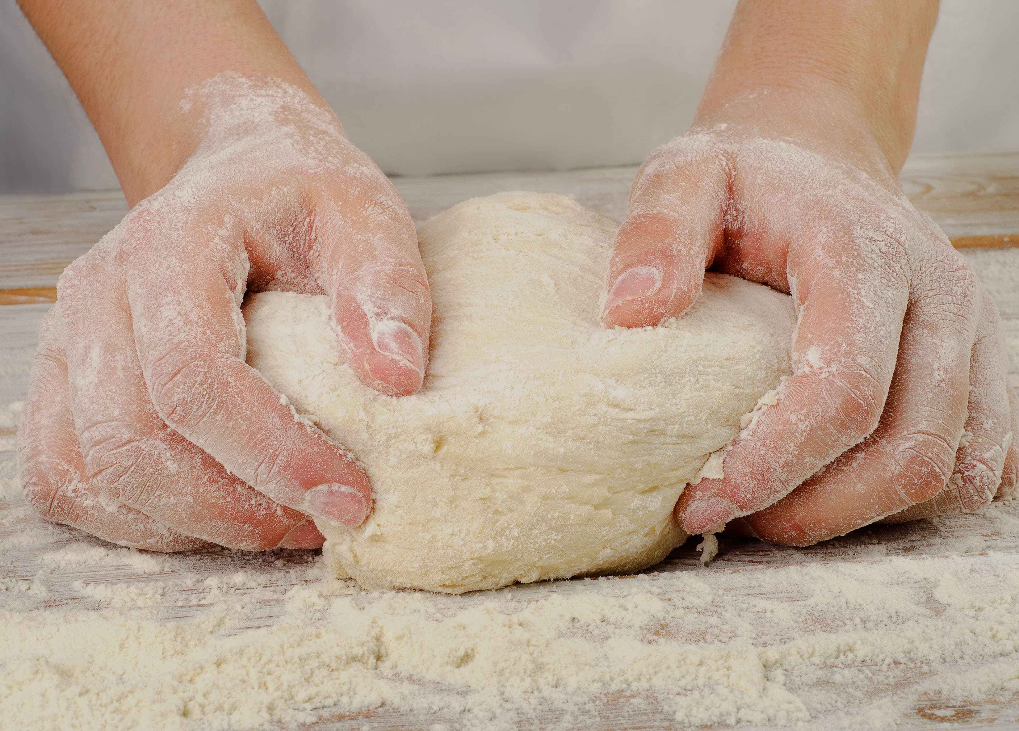 A pair of floured hands kneading dough on a wooden surface.